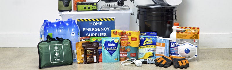 First aid kit, bottles of water, tinned and packaged food, gloves, charger, buckets, radio, mask, cleaning materials and a plastic box labelled 'Home emergency supplies'.