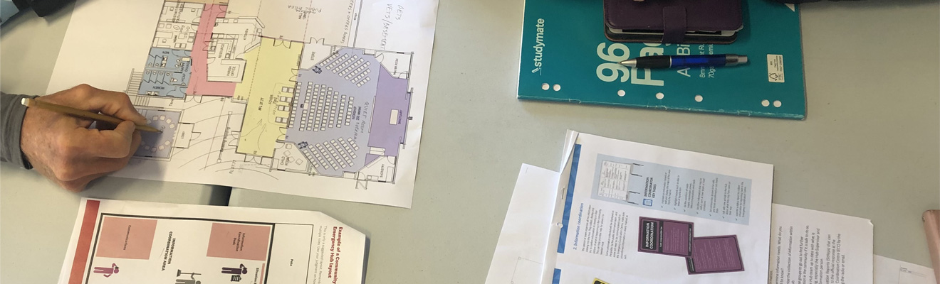 A table with emergency planning documents, a notepad and pen and a hand holding a pencil writing on a building plan. 